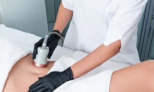 A person in a white coat uses a treatment device on a patient's abdomen while wearing black gloves. The patient is covered with a towel.