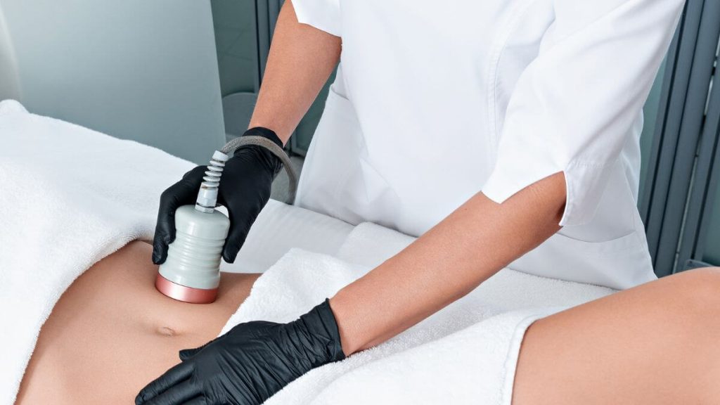 A person wearing gloves is using a device on a client's abdomen during a cosmetic treatment.